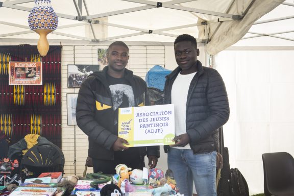 Deux hommes souriants tiennent une pancarte dans un marché avec divers objets en arrière-plan.
